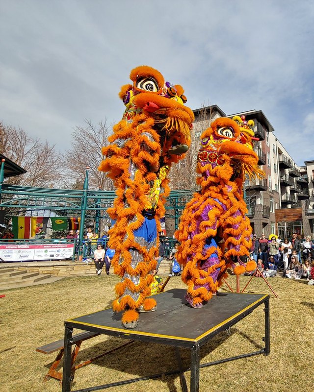 Two orange lion dancers perform on a raised platform before a crowd in a plaza with apartment buildings behind them at an Arvada Winterfest event.