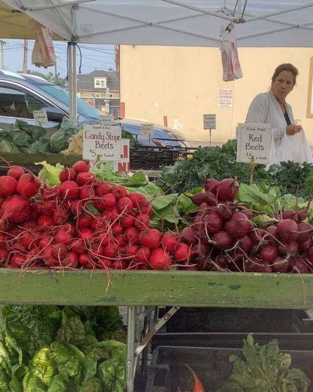 Red radishes and red beets on a market table under a white tent, with a vendor standing behind it at a farmers market.