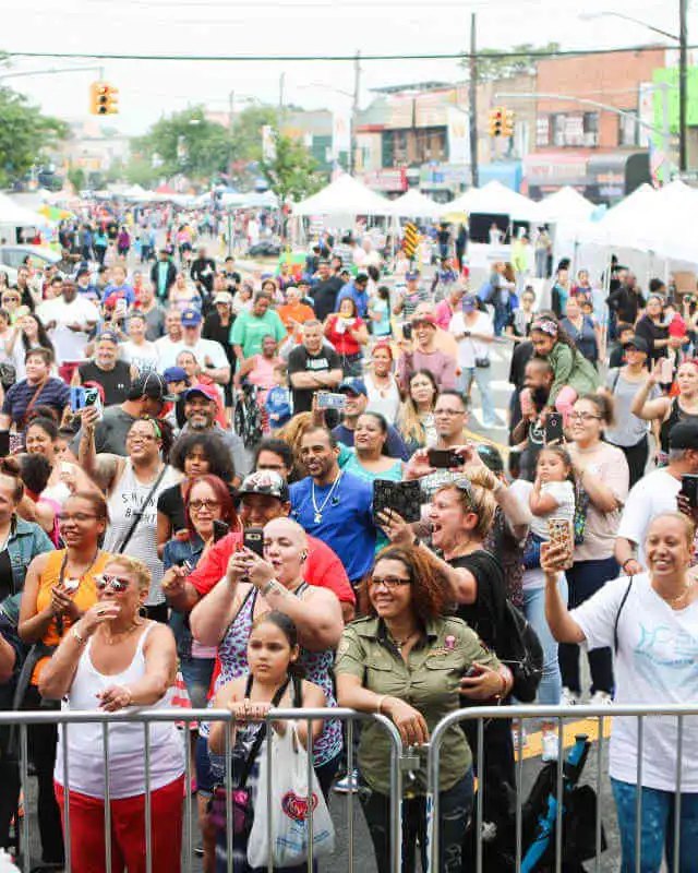 Large crowd gathered along a street beside white vendor tents at an outdoor Bronx fair.