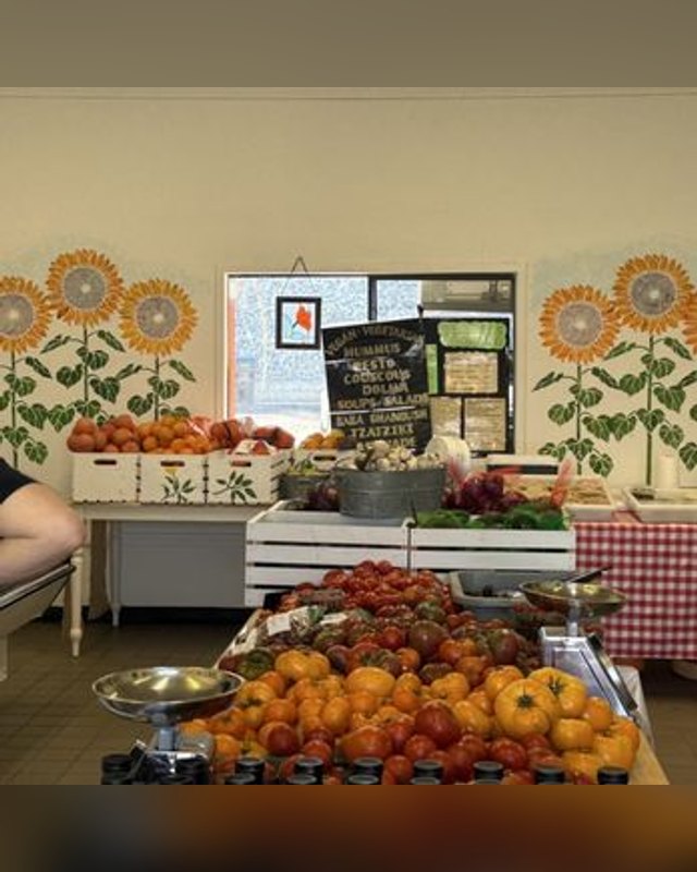 Produce stand with tomatoes and peppers under sunflower murals, with a chalkboard sign and a scale at a market booth.