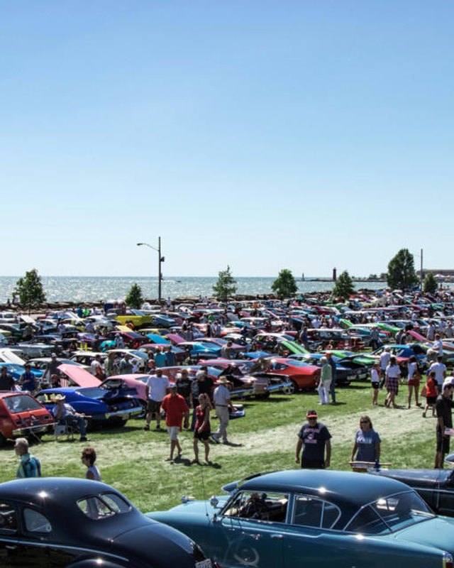 Rows of classic cars parked on a lakefront lawn with spectators walking among them during a car show.