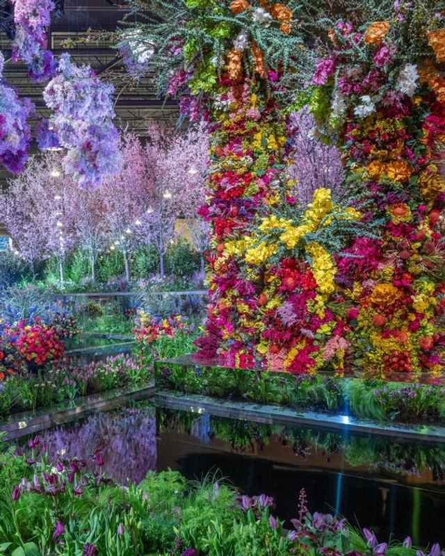 Flower show installation with a tall wall of colorful blooms beside a reflecting pool and blossom trees under lights.