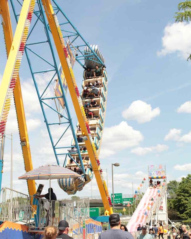 Fairgoers walking past a tall swinging thrill ride on a sunny midway