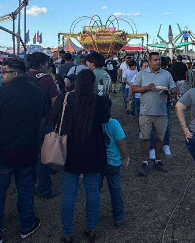 Visitors standing in a crowd near a carnival ride and a white tent at an outdoor fair, with a man holding food in the center.