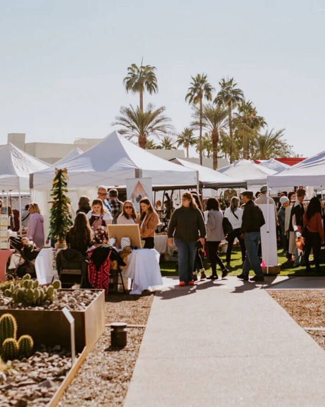 Shoppers browsing white market tents on a palm-lined walkway at the outdoor Merchantile Market