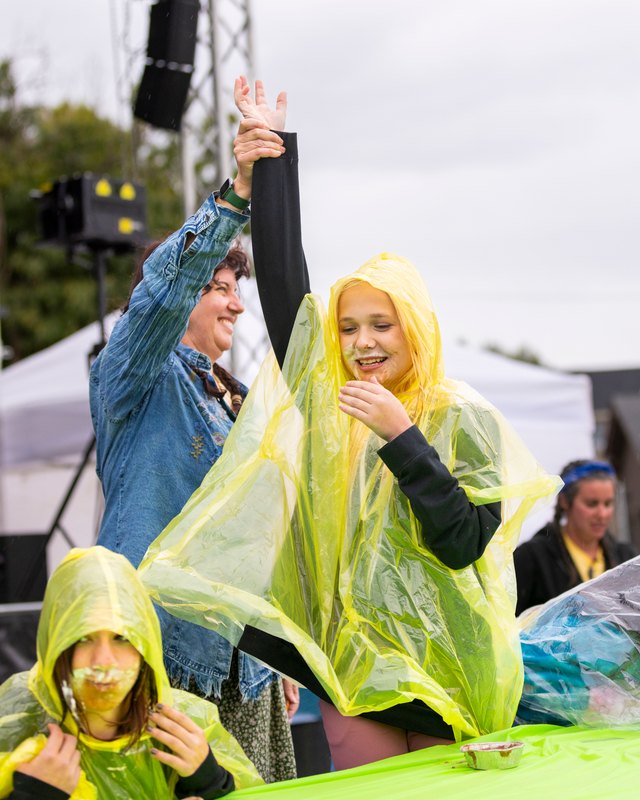 Kids in yellow rain ponchos at the Edgewater Hometown Fest pie eating contest, with an adult cheering beside them.
