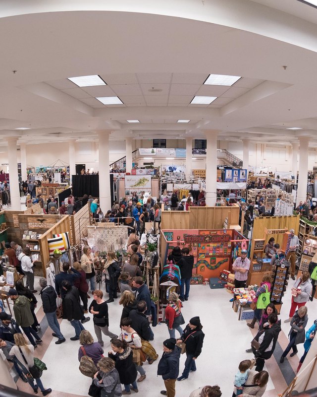 Wide view of a crowded indoor market with rows of vendor booths and shoppers under high ceilings.