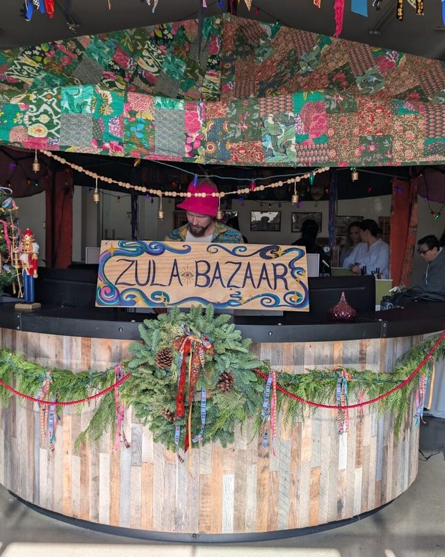 Zula Bazaar sign on a decorated wooden market booth with greenery and colorful fabric overhead