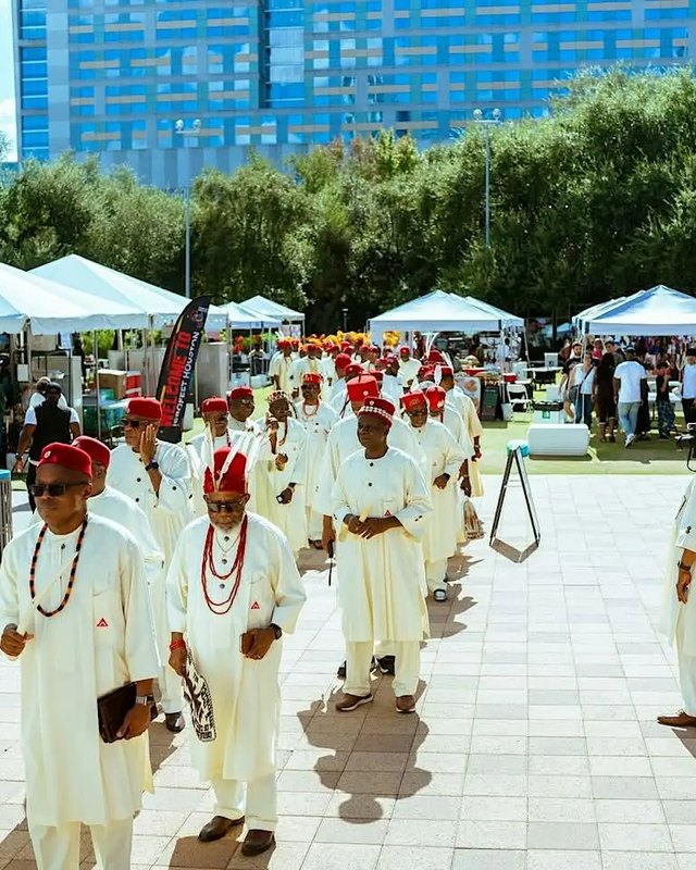 Men in white robes and red caps walk past vendor tents at an outdoor IGBOFEST festival in Houston.