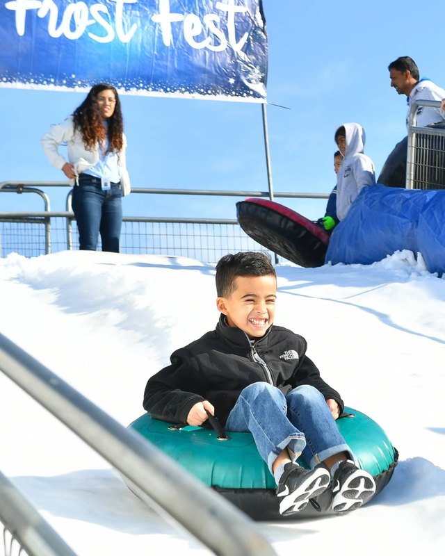 A smiling boy rides a snow tube down a slide at Frost Fest, with a Frost Fest banner and people behind him.