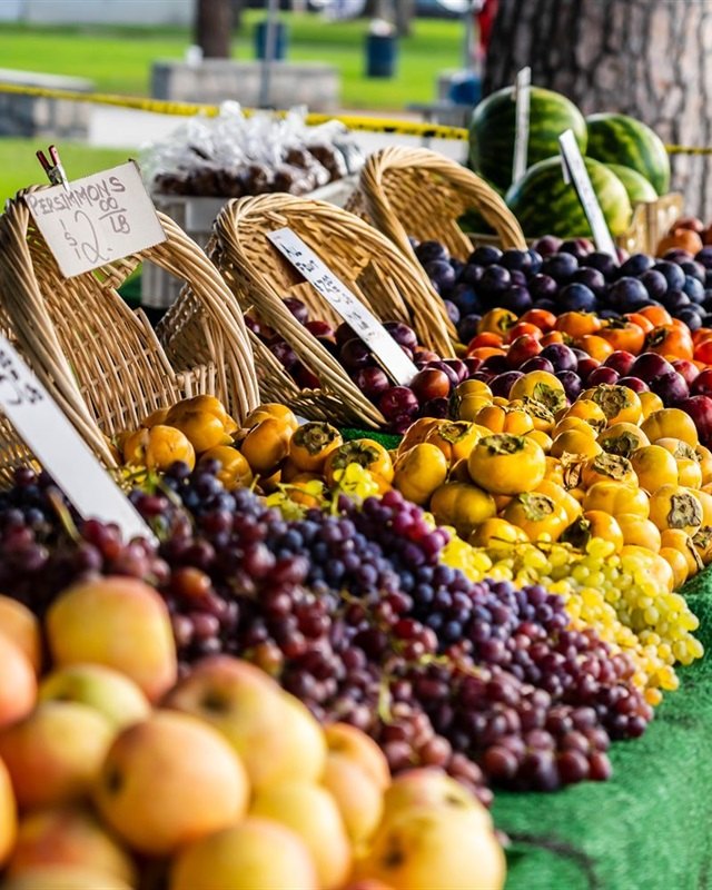 Piles of apples, grapes and colorful produce on a green-draped market table, with wicker baskets and price signs behind.