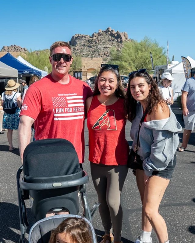 Three adults posing together at an outdoor market, with a stroller in front and white tents and a rocky hill behind them.