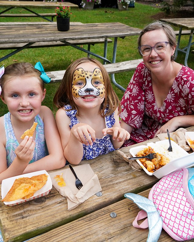 Three smiling people at a wooden picnic table with plates of food; a girl on the right has face paint of a leopard. Outdoors on grass.