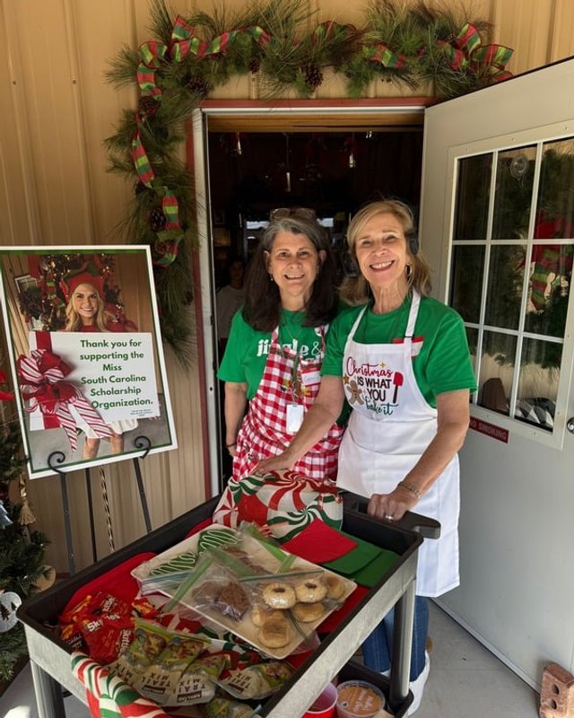 Two smiling volunteers in green shirts and red and white aprons serving baked goods from a cart under a festive garland at Tom's Den.