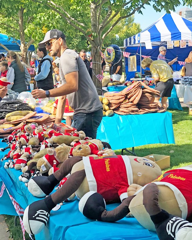 Shoppers at Potato Days booths under blue and white tents, with plush potato toys in red jerseys and baked-potato-style goods on display outdoors.