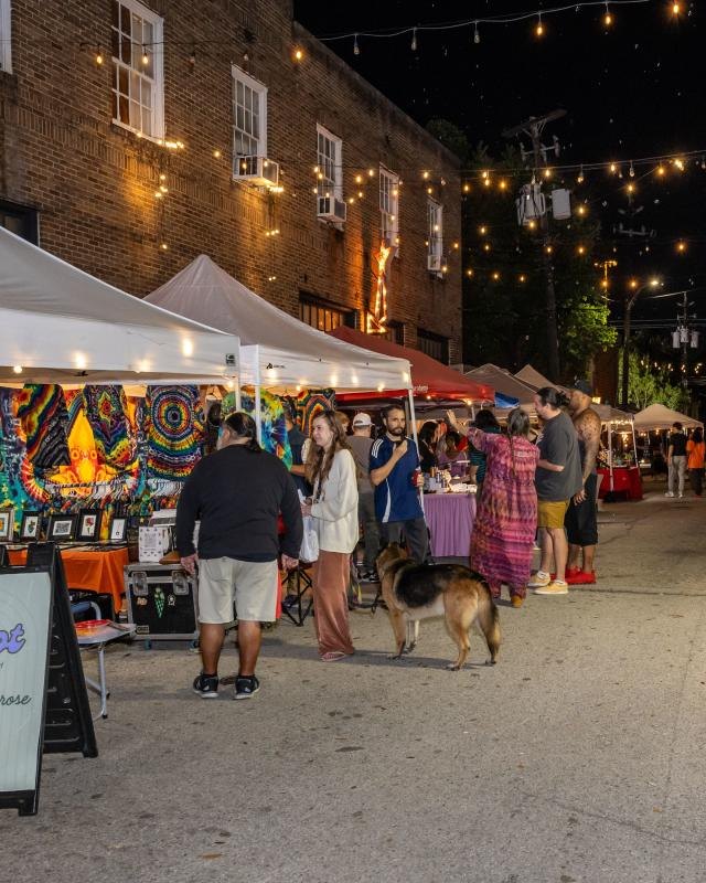 Night street market with white booth tents, string lights and shoppers browsing along a brick-lined street.
