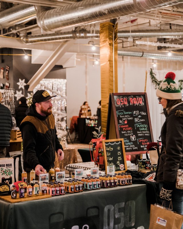 A vendor in a black cap stands behind a booth of bottled sauces and goods as a shopper in a Santa hat looks on at an indoor holiday bazaar.