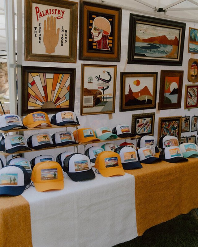 Hat and framed-art booth under a white tent at an outdoor vendor market, with mustard and white table covers.