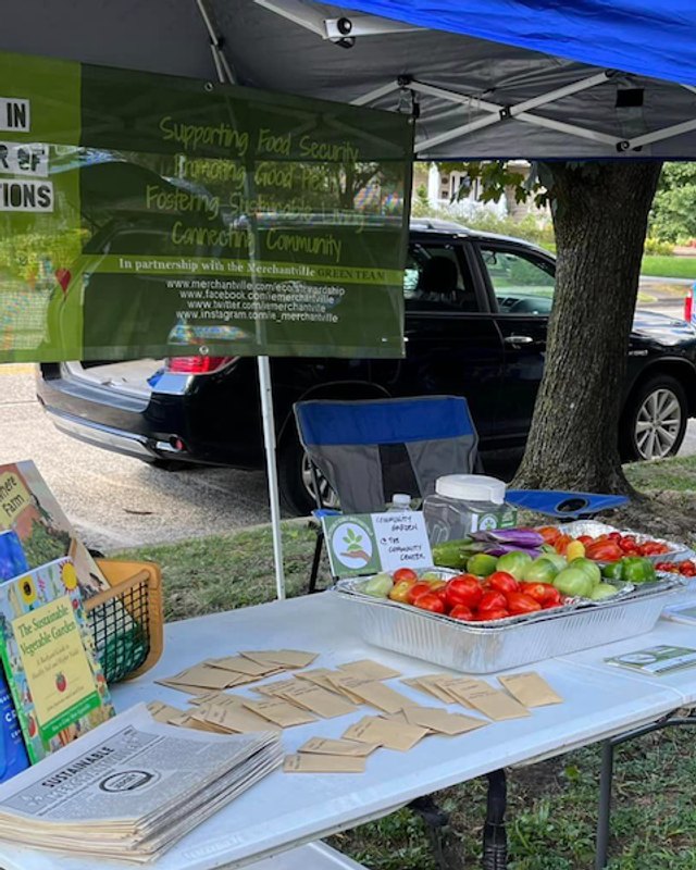 Produce and goods laid out on a table under tents at an outdoor market with parked cars nearby.