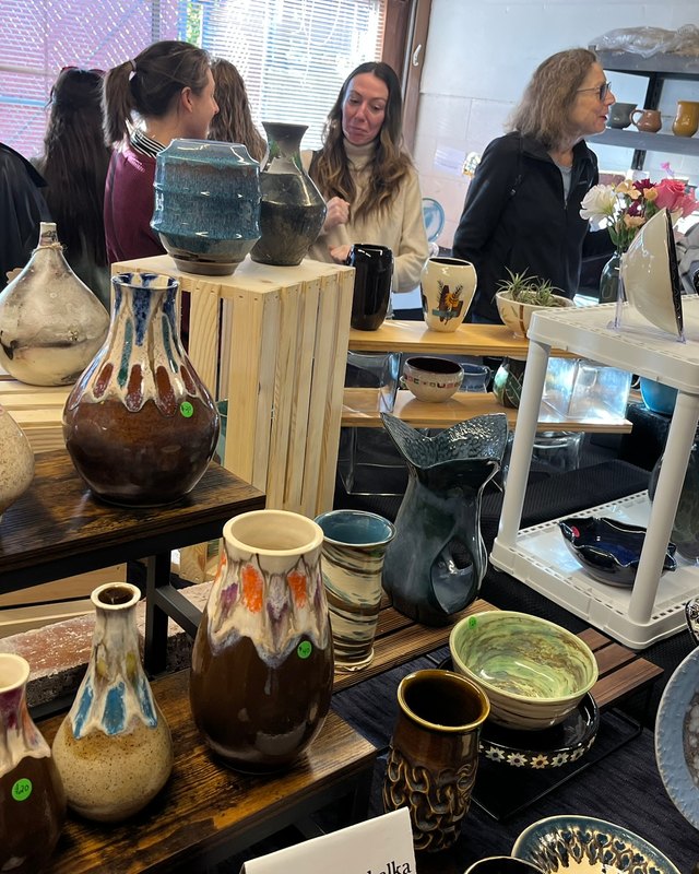 Shoppers gather around a display of glazed pottery vases and bowls on tables at a pottery market booth.