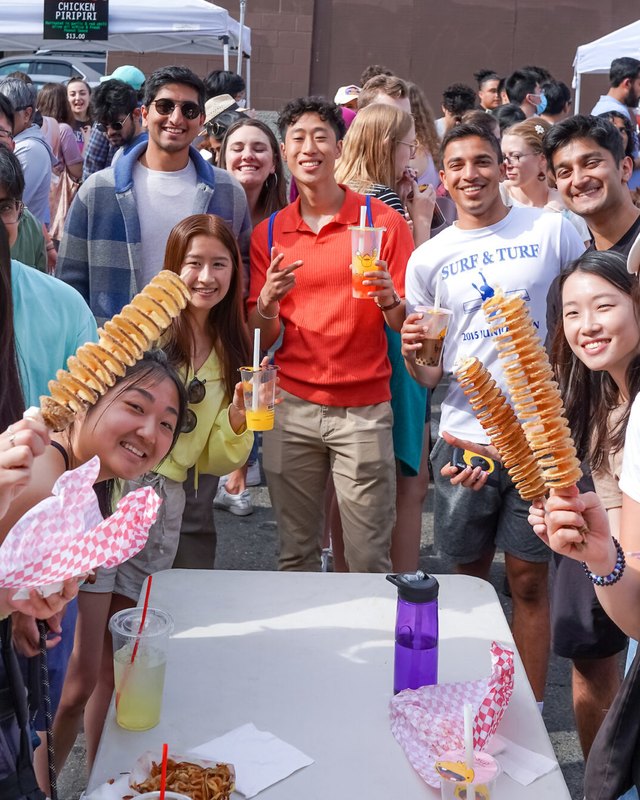 Festival-goers crowd around a food booth at the U District Street Fair, holding snacks and smiling.