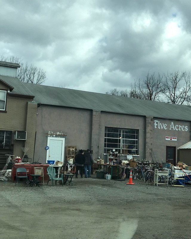 Outdoor vendor tables with goods and shoppers in front of the Five Acres building under a cloudy sky.