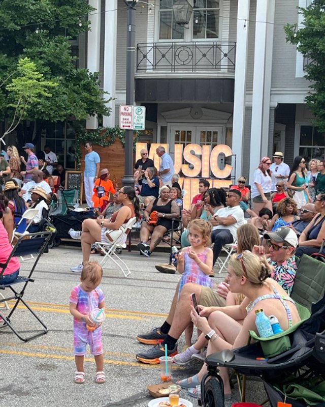Crowd of people seated and standing along a Clayton street during the music festival, with a MUSIC sign behind them.