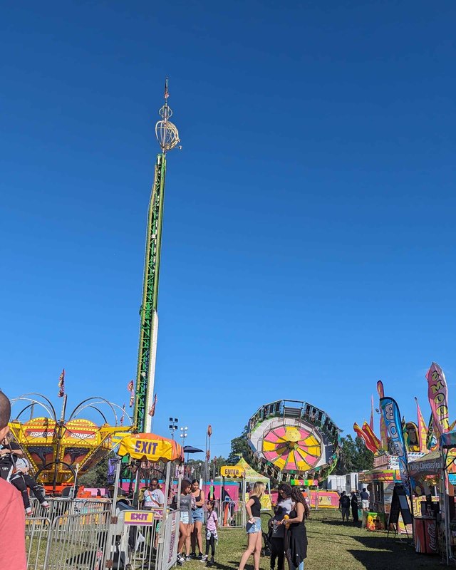 Tall tower ride and a spinning ride over a fenced midway with food booths and visitors at the Hernando County Fair, under a clear blue sky.