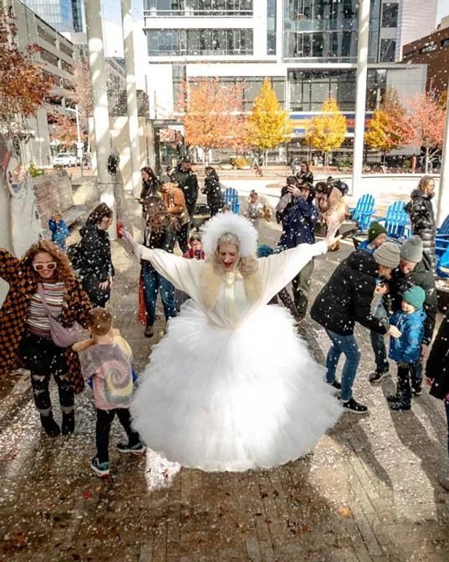 Crowd gathered in an outdoor plaza at Snow Day Village, with a performer in a white costume dancing among them.
