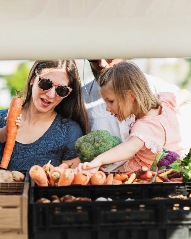 A smiling mother and two children with fresh produce at a market stall.