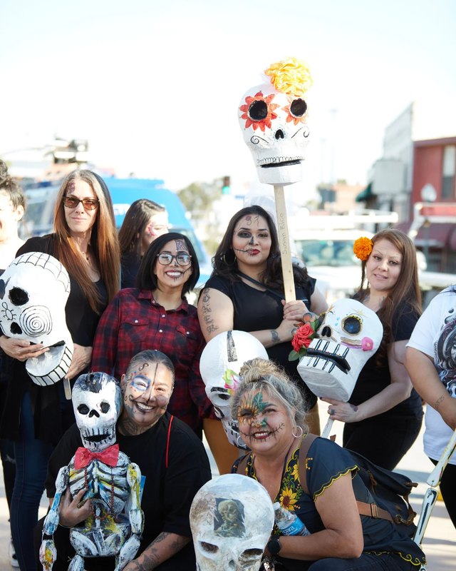 Group of people posing with large skull masks and calaveras on a city street, some in black clothing, at the Self Help Graphics Día de los Muertos event.