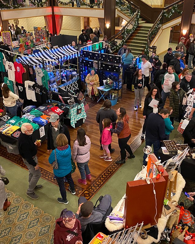 Shoppers browse vendor booths, including a striped-awning table of shirts, in a crowded decorated ballroom at a holiday market.