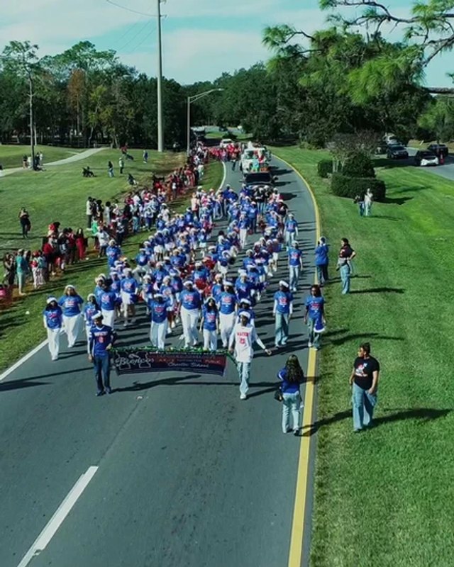 Marching parade participants and spectators on a road lined with trees in Poinciana