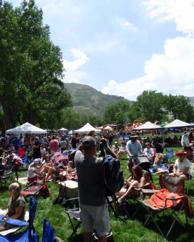 Crowd gathered on a grassy lawn with white vendor tents and mountains behind at the 4th of July Festival in Golden.