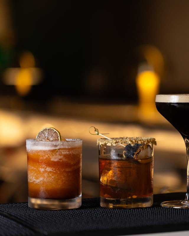 Three cocktails with a coupe glass on a dark bar counter, warmly lit.