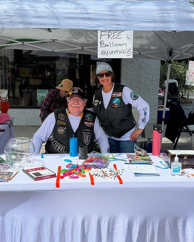 Two people smile behind a white-draped table under a white tent, with a "FREE Balloon Animals" sign and brochures on the table at a street event.