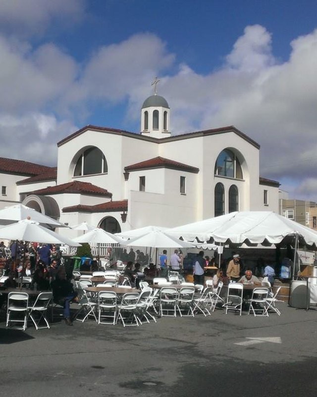 White-tented festival booths and people seated at tables in front of the white cathedral buildings of Annunciation Cathedral.