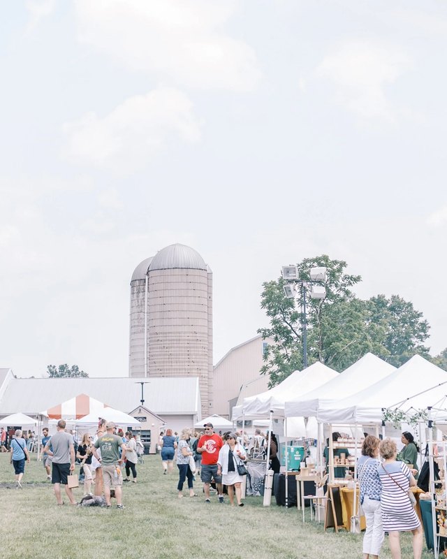 Shoppers walking among white booth tents on a grassy field with silos behind at the Bishop Estate market.