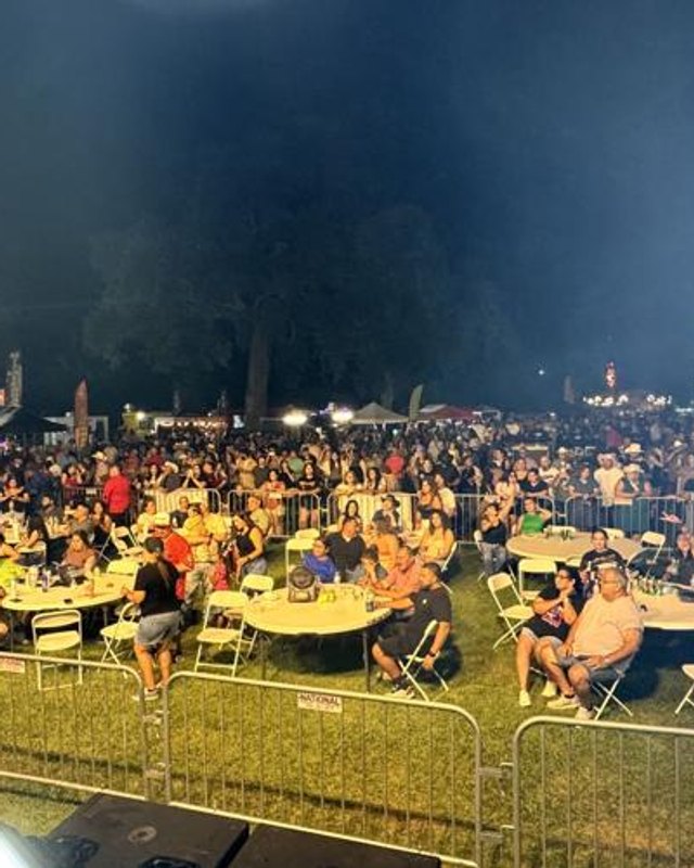Night crowd seated at tables and standing behind metal barricades at an outdoor festival in a park, with string lights in the background.