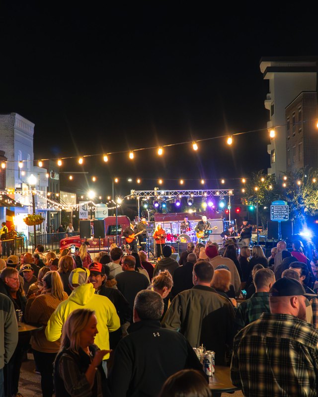 Evening street crowd gathered under string lights, facing a lit stage with a band on a West Columbia street.