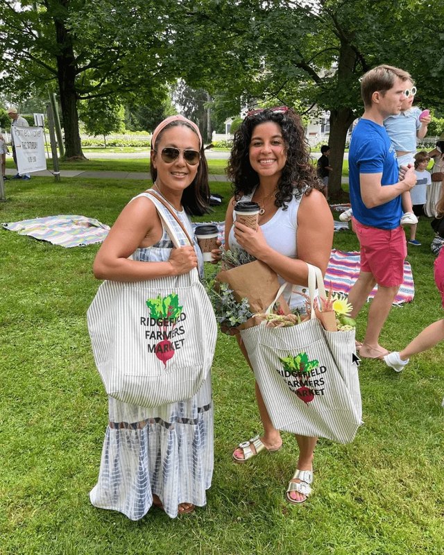 Two shoppers carrying Ridgefield Farmers Market tote bags on a grassy park lawn during the market.