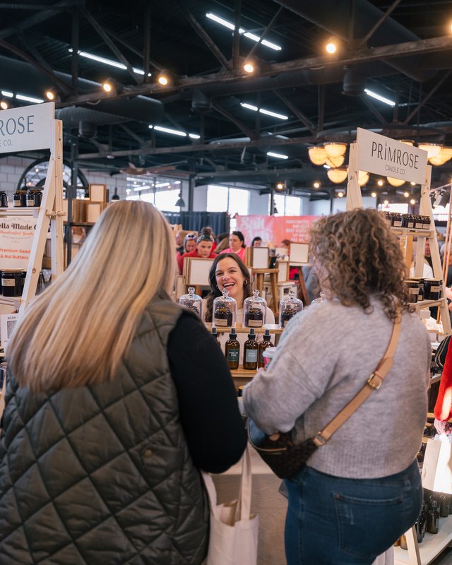 Two shoppers at a market booth with bottled products and a Primrose sign, under string lights in an indoor market hall.