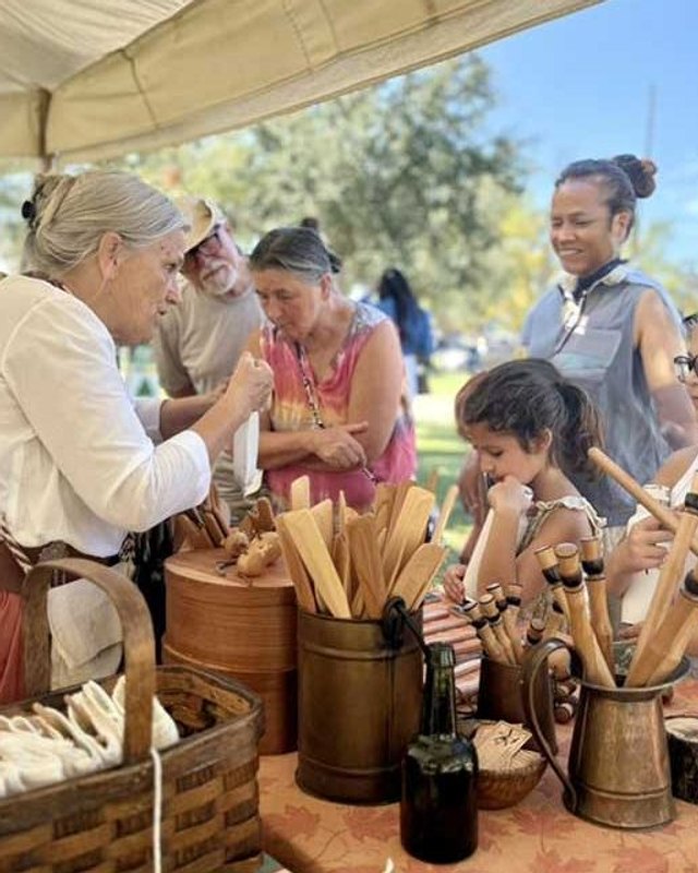 Costumed interpreters and children gathered at a historic pioneer-style table with baskets and wooden tools.