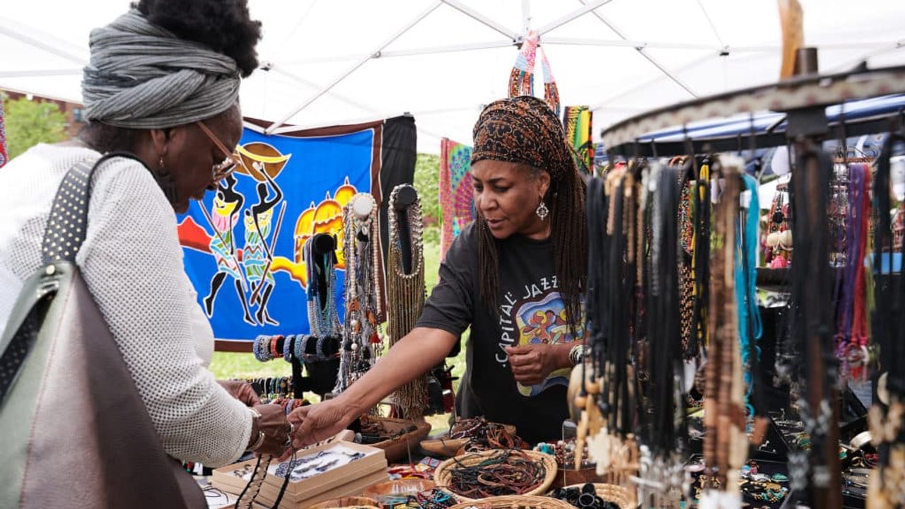 Shoppers and vendors at an artisan market booth with colorful textiles and jewelry under a white tent.