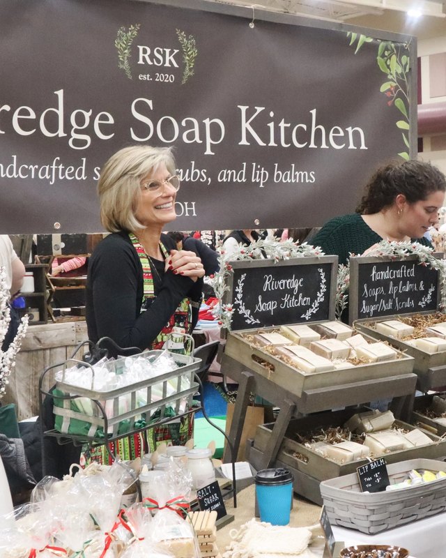 Smiling vendor at the Riveredge Soap Kitchen booth at a holiday craft show, with soap bars and gift bags on display.