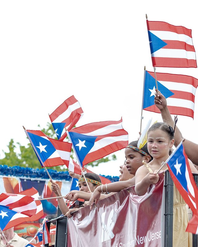 Young girls waving Puerto Rican flags from a parade float, with a pink banner below.