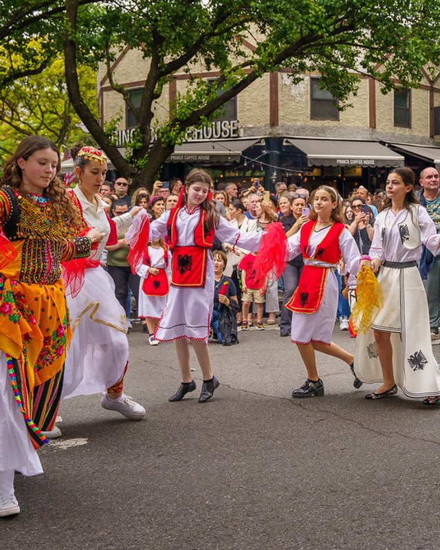 Costumed dancers and crowds parading down a tree-lined street at an Albanian street event.