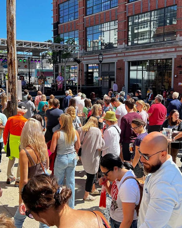 Crowd of festival-goers packed along a sunny street near brick buildings at the Summer Soulstice Music Festival.