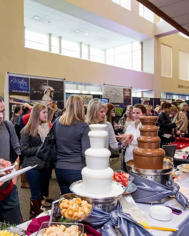 Busy expo hall with attendees browsing vendor booths and a chocolate fountain display at the Ultimate Wedding Expo.