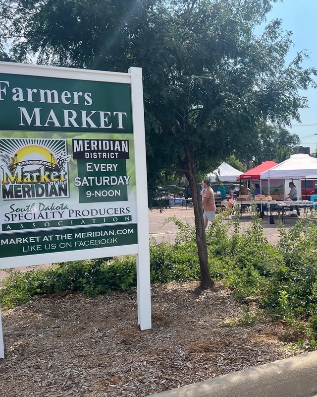 Farmers Market sign for Market at the Meridian with white market tents and vehicles behind it in Yankton.